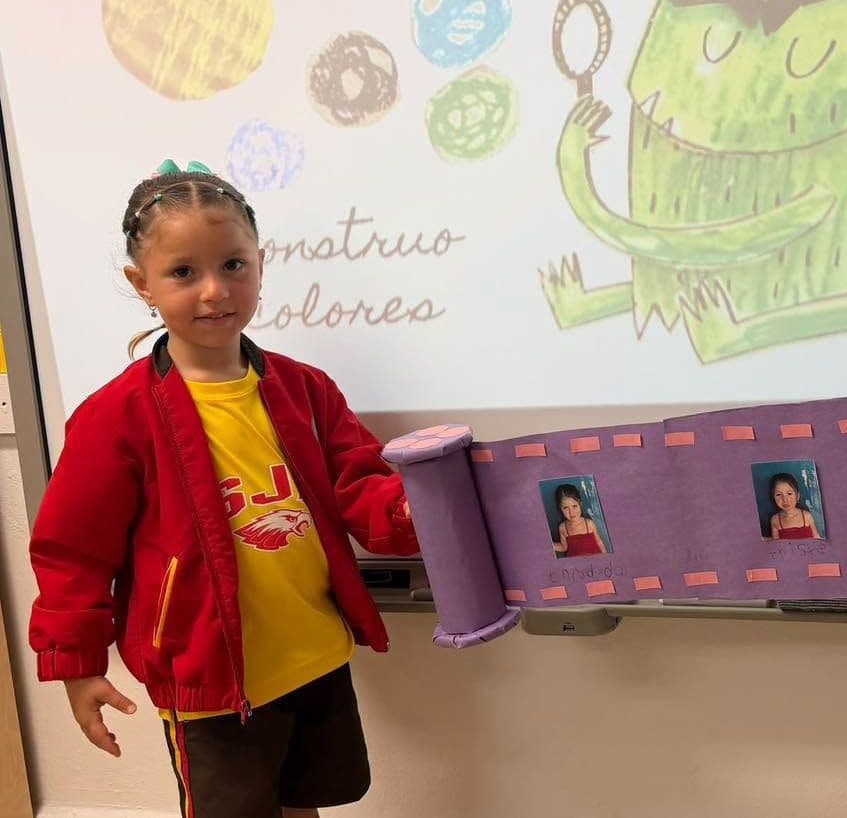 Niña de preescolar con chaqueta roja junto a su trabajo en la pared